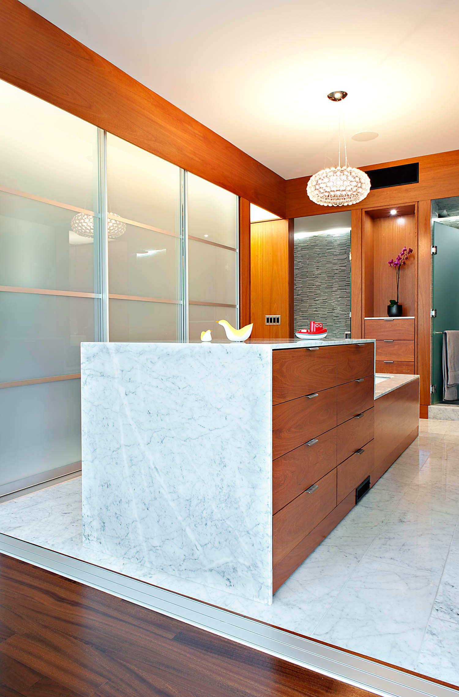 浴室的远景，突出了大理石地板和浴缸的环绕，配有嵌入式木抽屉存储。烟熏玻璃隐私墙的功能在左边。