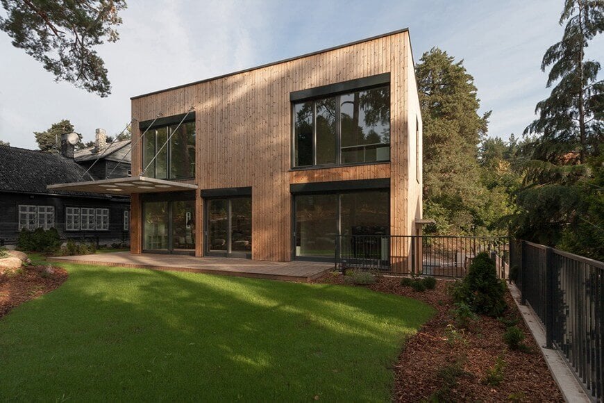 一间带有木制外观和玻璃窗和门的Modish的房子。房子提供甲板和大型草坪和花园区。