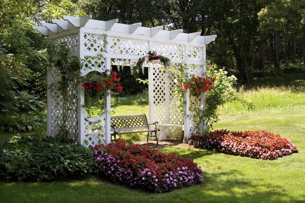 这个白色的藤架坐落在一个小花园露台上，上面装饰着吊篮、攀缘藤蔓和厚厚的花坛。藤架的结构由棚架构成。