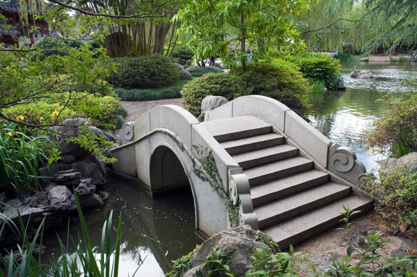 一座华丽的混凝土桥横跨一条宽而浅且缓慢流动的小溪。这座桥连接了一个广阔、郁郁葱葱的花园的两个部分。
